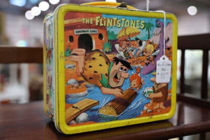 Vintage 1962 Flintstones metal lunchbox featuring Fred Flintstone and other Hanna-Barbera characters at a prehistoric resort scene, displayed at the Chicagoland Coin-Op Show.