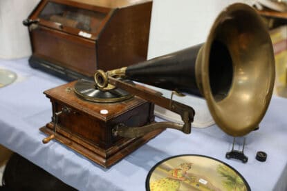 Antique external horn phonograph with hand crank, brass tonearm, and wooden cabinet, on display with an illustrated vintage record at the Chicagoland Coin-Op Show.