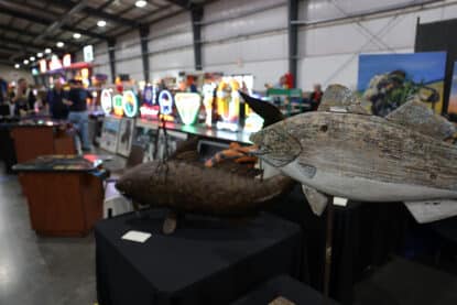 Folk art wooden fish sculptures displayed alongside a wall of vintage neon beer and diner signs at the Chicagoland Coin-Op Show.