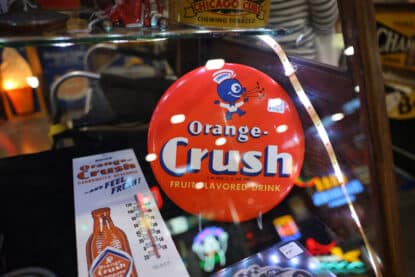 Vintage Orange Crush round soda sign featuring mascot Crushy, along with a branded thermometer, on display at the Chicagoland Coin-Op Show.