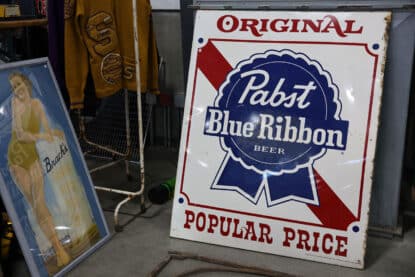 Vintage enamel sign advertising Pabst Blue Ribbon beer with red and blue graphics and the slogan "Original - Popular Price", alongside other retro memorabilia.