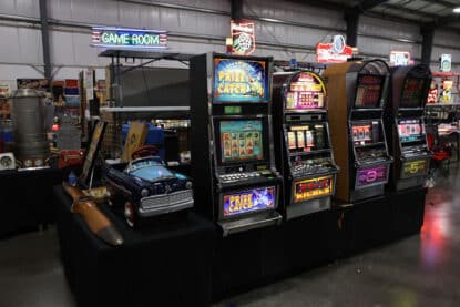 Row of modern and vintage slot machines under a neon 'Game Room' sign.