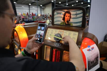 Ed Owens measuring the grill of a Rock-Ola Mystic Music jukebox while photographing the process with a smartphone at a show booth.