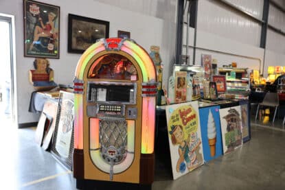 A glowing Rowe AMI jukebox surrounded by vintage advertising signs and collectibles at an antique show.