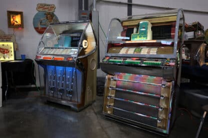 Two vintage Seeburg jukeboxes, a HF100R and V200, illuminated in blue and multicolor lights, displayed side by side at a retro show.