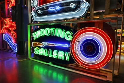Vintage style neon sign reading "Shooting Gallery" with a glowing green rifle, red bullseye target, and vibrant lighting effects, displayed at a Chicagoland antique sign show.