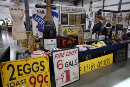 Assortment of vintage Americana signs including Fire Chief gas price, EAT neon, and Bon Ton mannequin at antique show.