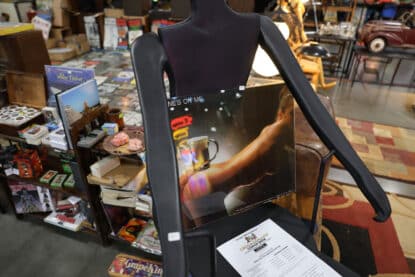 Vintage beer poster featuring a muscular back with a beer mug balanced on the shoulder, displayed on a modern mannequin chair at the Chicagoland Coin-Op Show.