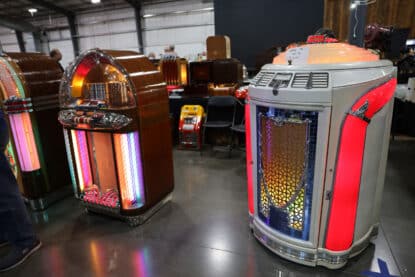 Two vintage jukeboxes, including a Seeburg and Wurlitzer, displayed at a collector event, both fully restored with vibrant illumination and chrome detailing.