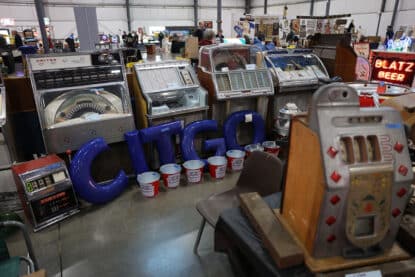 A collection of vintage jukeboxes, a slot machine, large CITGO letters, and Budweiser beer buckets at an antique Americana show.