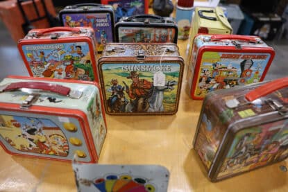 Assorted vintage metal lunchboxes featuring Gunsmoke, Disney Express, The Flintstones, Looney Tunes, Super Heroes, and more on a wooden display table.