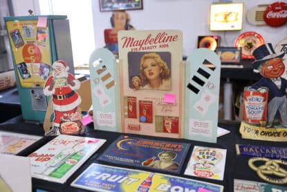 Vintage Maybelline eye and beauty aids display surrounded by colorful advertising signs for Heinz soup, Crescent soda, Bubble Up, and more.
