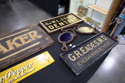 Assortment of vintage medical and dental office signs including 'DR. W.L. COOK DENTIST' and 'G.R. EADES M.D. EYE EAR NOSE & THROAT' alongside antique optometry lenses and enamel signage.