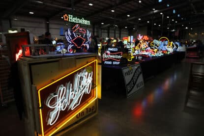 Row of colorful vintage neon beer signs including Schlitz, Budweiser, Heineken, and Pabst, displayed on tables at an antique show.