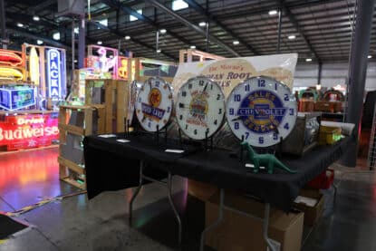 Vintage advertising clocks for OK Used Cars, Pep Boys Motor Oil, and Chevrolet Sales & Service displayed on a table, surrounded by neon signs at a collectibles show.
