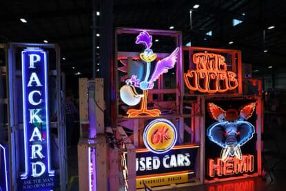 Collection of vintage neon signs including Packard, Road Runner, OK Used Cars, The Judge, and HEMI displayed on wooden frames at a collector show.