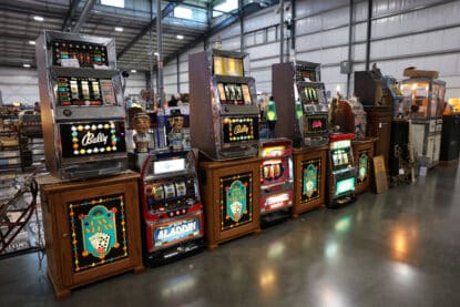 Lineup of vintage Bally slot machines and other retro casino games on themed wooden cabinets at an antique show.