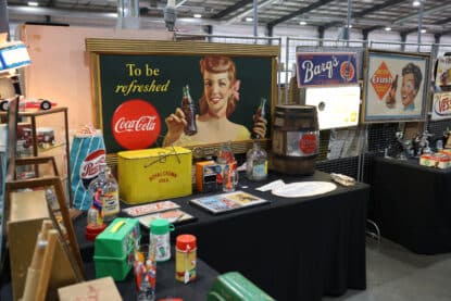 Vintage soda memorabilia display featuring Coca-Cola, Pepsi, Barq's, Crush, and Royal Crown Cola signs, bottles, and promotional items on a black table at a collector's show.