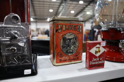 Close-up of a vintage Tiger Chewing Tobacco tin and a sealed Jack o’ Diamonds cigarette pack on a collectible vendor table, flanked by antique vending machines.