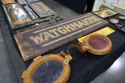 Collection of vintage signs including a large wooden Watchmaker sign, District Court plaque, and oversized antique optometry lenses.