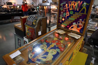 Vintage Williams Perky pinball machine with colorful artwork and illuminated playfield, next to an antique coin-operated slot machine at an antiques show.